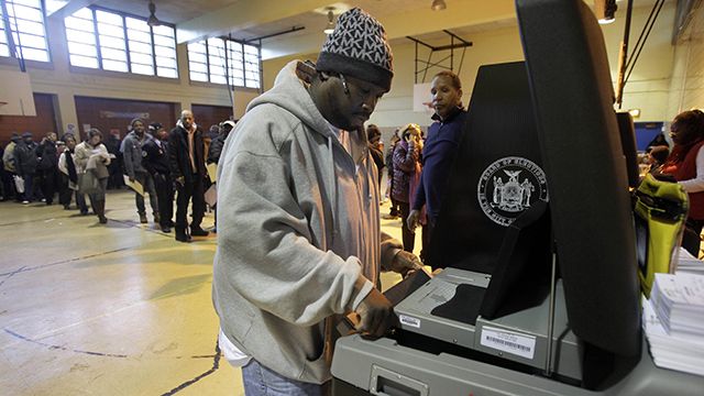 Poll worker Eric Carr, background center, watches a technician for the New York City Board of Elections clear a paper jam in a ballot scanner as voters wait to scan their ballots, at a school in New York's Harlem neighborhood, Nov. 6, 2012. (AP Photo/Richard Drew)