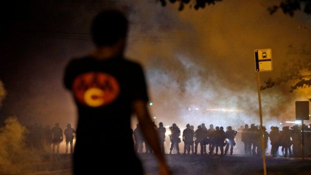 A man watches as police walk through a cloud of smoke during a clash with protesters Wednesday, Aug. 13, 2014, in Ferguson, Mo. Protests in the St. Louis suburb rocked by racial unrest since a white police officer shot an unarmed black teenager to death turned violent Wednesday night, with people lobbing Molotov cocktails at police who responded with smoke bombs and tear gas to disperse the crowd. (AP Photo/Jeff Roberson)